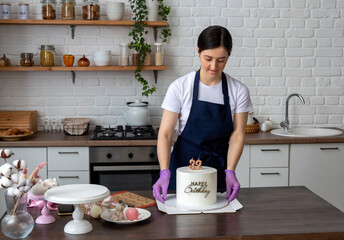 Woman in an apron holds white cake with the inscription Happy birthday and the numbers 32 in her hands and puts it on cardboard box. Packaging for delivery. Selective focus.