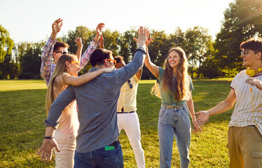 Group of happy young people have fun together while walking in park on warm summer evening. Cheerful excited multiethnic friends laugh out loud and give each other high five. - obrazy, fototapety, plakaty
