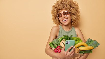 Glad woman uses mobile phon for ordering products online carries fresh vegetables and fruits bought...