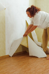 Side view of young plump woman with long curly red hair hair wearing white T-shirt, beige trousers, leaning down to tear off yellow wallpaper from wall. Home redecoration, interior change. Vertical.