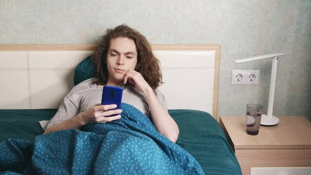 A young man sits reclining in bed and looks at the phone, then smiles a little. There is a glass of water and a lamp next to it. Slow motion 4k footage