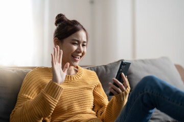 Happy young hispanic asian teen girl sit on sofa at home holding phone looking at screen waving hand video calling distance friend online in mobile chat app using smartphone videochat application.