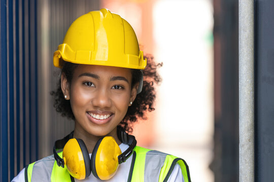 Young African American trainee in safety suit, yellow hard hat and earmuffmuff stand smiling next to big container in a shipyard. African curly hair female worker in smile. Female loader with earmuff
