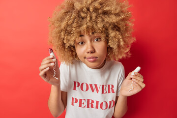 Menstruation gynecology and hygiene concept. Young curly haired European woman holds two tampons chooses best absorbency product wears white t shirt with inscription isolated over red background