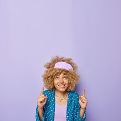 Vertical shot of positive young woman with curly hair wears sleepmask and nightwear points index fingers above smiles gladfully isolated over purple background. Advertising product for sleeping