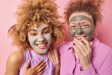 Two positive young women with curly hair apply beauty masks for skin treatment giggles happily stand next to each other isolated over pink background. Facial procedures and rejuvenation concept