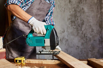 Female Carpenter sawing wood with an electric jigsaw. Woman in apron doing some carpentry work in...
