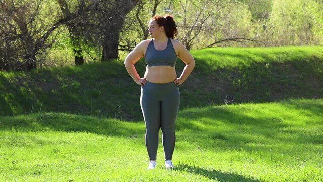 Young overweight woman in fitness suit turning neck side to side standing on green lawn in park on sunny day, nature background. Healthy lifestyle, exercise and fitness outdoors. Sports activity.