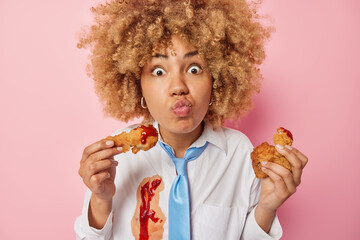 Surprised European woman with curly bushy hair eats tasty nuggets with ketchup keeps lips folded...