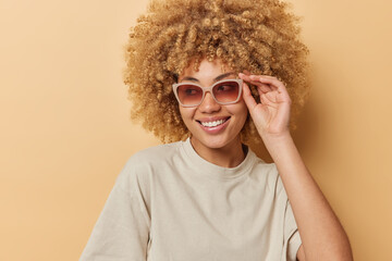 Indoor shot of happy curly haired young woman wears sunglasses and casual t shirt looks gladfully away has good mood isolated over beige background. Positive human emotions and style concept