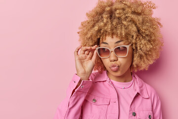 Photo of good looking young European woman with curly hair keeps lips folded wears sunglasses and jacket has stylish look isolated over pink background blank space for your advertising content