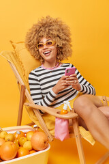 Vertical shot of cheerful young woman wears sunglasses and striped jumper holds mobile phone...