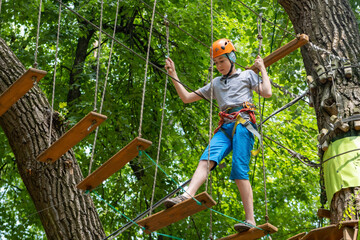 Rope park. A boy teenager in a helmet walks on suspended rope ladders. Carabiners and safety straps. Safety. Summer activity. Sport. Children's playground in nature in the forest. © Real_life