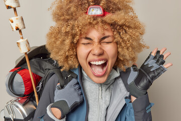 Emotional curly haired woman exclaims loudly wears headlamp jacket and gloves carries heavy backpack full of necessary things has adventure travel isolated over grey background. Camping and hiking © WHstudio Leushin N