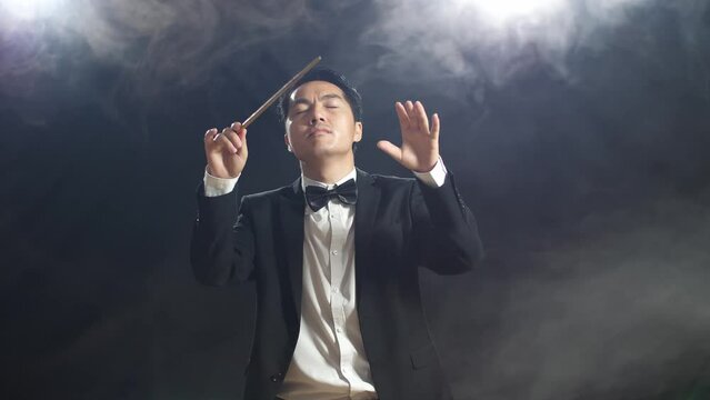 Asian Conductor Man Holding A Baton Closing His Eyes And Showing Gesture In The Black Studio With Fog 

