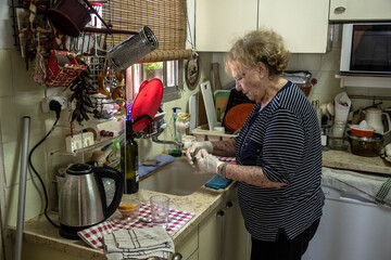 Elderly woman washes in her kitchen with rubber gloves. - obrazy, fototapety, plakaty