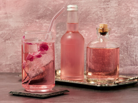 A glass of pink gin and tonic, a bottle of rose lemonade and a bottle of pink gin on a light pink background