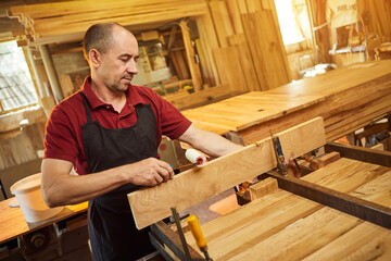 Portrait of a senior carpenter in uniform gluing wooden bars with hand pressures at the carpentry manufacturing