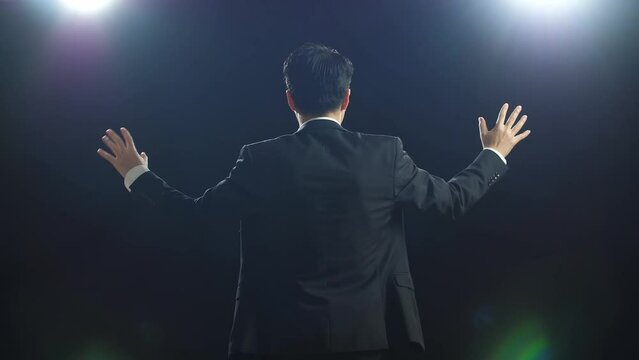 Back View Of Asian Speaker Man In Business Suit Showing Index Fingers Up While Speaking In The Black Screen Studio
