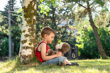 A happy in a red T-shirt boy hugs  a Teddy bear while sitting with his back to a tree
