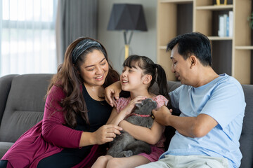 Happy diverse asian family couple with child daughter playing with pet cat on sofa in living room...