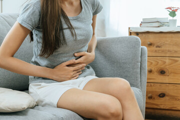 asian young woman suffering stomach ache sitting on couch in living room at home, people painful...