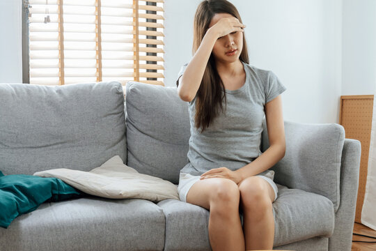 asian young woman touching forehead having headache migraine and upset with problem feel stressed sitting on couch in living room at home, people painful, depression, medical and health care concept