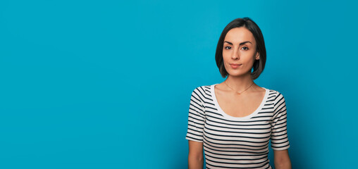 Banner photo of a beautiful confident young brunette woman is posing in a striped t-shirt and looking on camera isolated on blue background