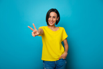 Photo of happy beautiful young girl with stylish hairstyle in yellow t-shirt while she shows victory sign and isolated on blue background in studio