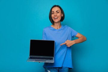 Attractive smiling brunette woman is presenting the modern laptop with blank screen and pointing on...
