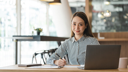 Young Asian businesswoman working on Laptop at desk, looking to a camera.