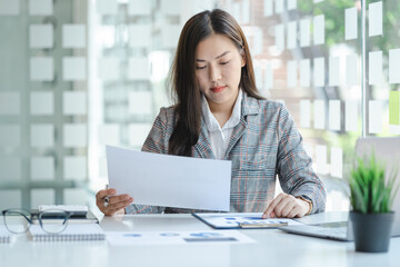 Smiling asian business woman sitting at office desk with computer, holding financial documents,...
