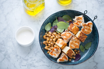 Halloumi and chicken kebabs with roasted chickpeas in a blue bowl, elevated view on a light-grey marble background, horizontal shot