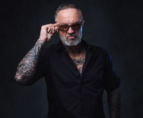 Studio shot of brutal aged man with sunglasses and tattooed body against dark background.