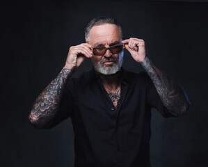 Studio shot of brutal aged man with sunglasses and tattooed body against dark background.