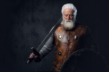 Portrait of gray haired medieval knight with long beard dressed in chain mail holding sword and...