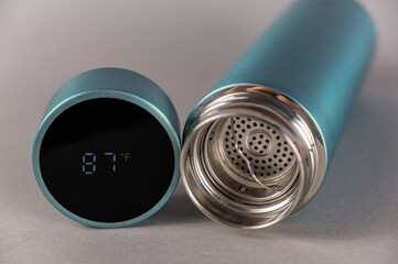 Thermos with open lid and teapot on gray background. Cylindrical lid with LED digital display. Temperature reading 87 degrees Fahrenheit. Vacuum flask of turquoise color.