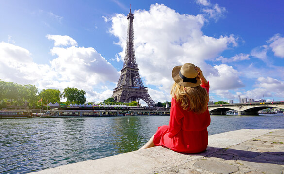 Young traveler woman in red dress and hat sitting on the quay of Seine River looking at Eiffel Tower, famous landmark and travel destination in Paris.