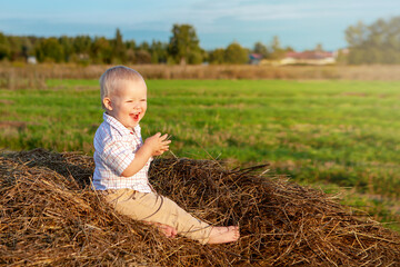 Naklejka na ściany i meble An emotional portrait of a happy little boy sitting on a haystack in a field and enjoying a warm summer day