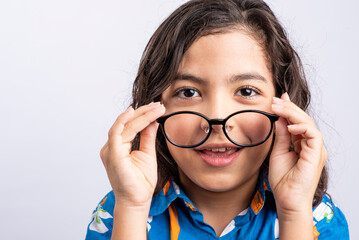 Image of a small boy in a blue shirt, looking at the camera while putting on his glasses, with a white background. © STOCKEROcr