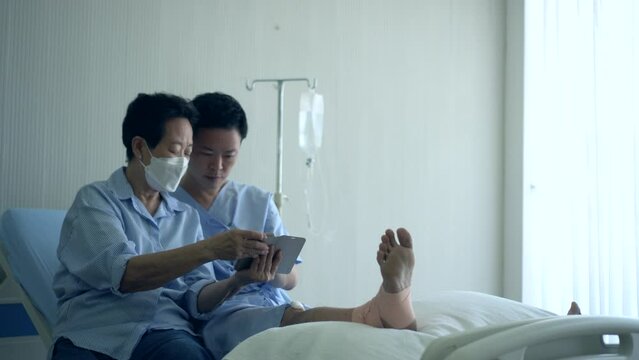 Asian mother and son who indured leg in hospital bed sharing conversation about social media on smartphone