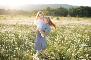 Cute beautiful young woman holding hands and play with little child girl walk in chamomile field with blooming flowers over nature background. Family lifestyle concept. Mom and baby daughter in grass