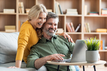 Happy middle aged couple websurfing on laptop together while relaxing at home