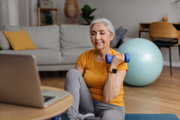 Domestic online training. Senior woman doing exercises with dumbbells in front of laptop, following...