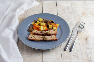 Sliced roasted pork meat in plate with vegetables on wooden table with rosemary and peppers.