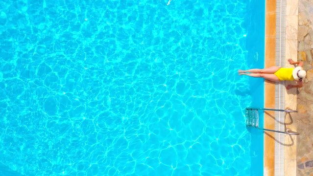 Aerial view of a woman in yellow swimsuit and a hat sits on the edge of the pool
