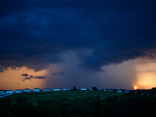 Dark clouds and turbulent sky with lightning and storm approaching and light at sunset over suburb houses on a green hilltop.