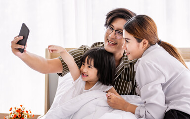 Asian warm family, father, mother, daughter playing, taking selfie photo together on bed in comfortable bedroom at home with love and fun, smiling with happiness. Lifestyle, Education and Kid Concept.
