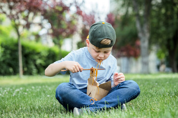 Child eating stir fried udon noodles sitting on the grass. Dirty stain of sauce on his t-shirt....
