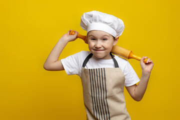 A little boy in a chef's uniform holds a rolling pin, isolated on a yellow background. Boy chef. Concept Of Cooking Process. Copy space